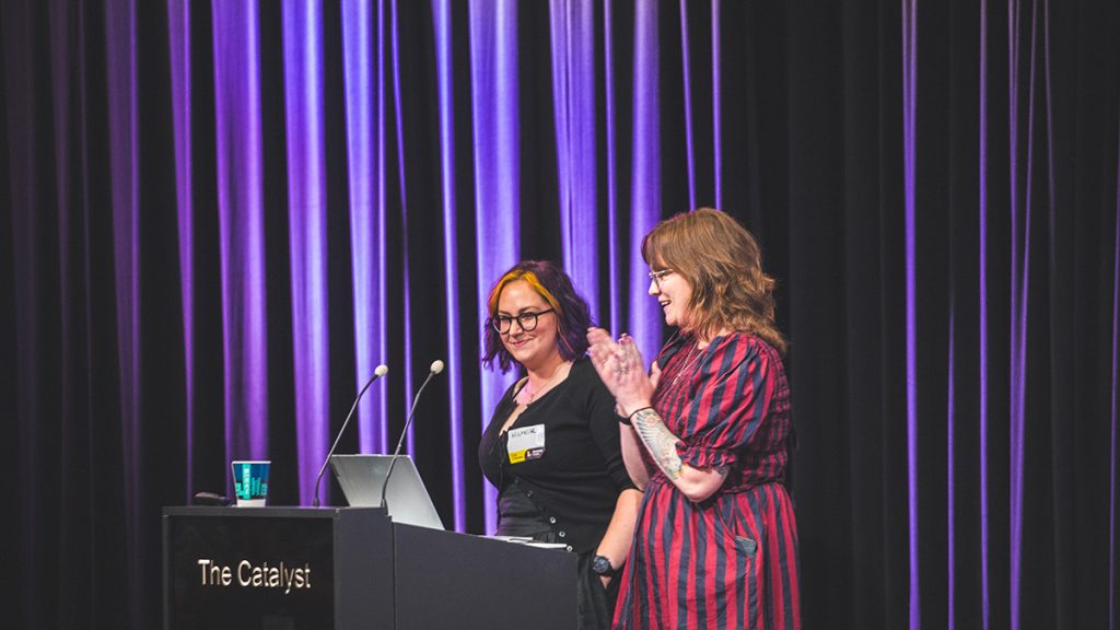 Michelle and Ellen on stage, in front of a subtle purple light. They are speaking at a lectern, Ellen is clapping.