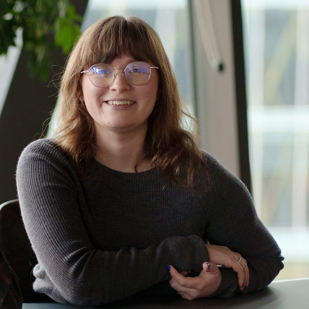 Ellen is a white woman with light brown hair and glasses, she's sitting at a table, with her arms crossed and smiling.